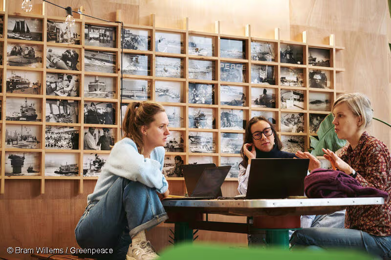Drie vrouwen werken samen aan laptops aan een tafel, met een wand vol historische zwart-wit foto's op de achtergrond.