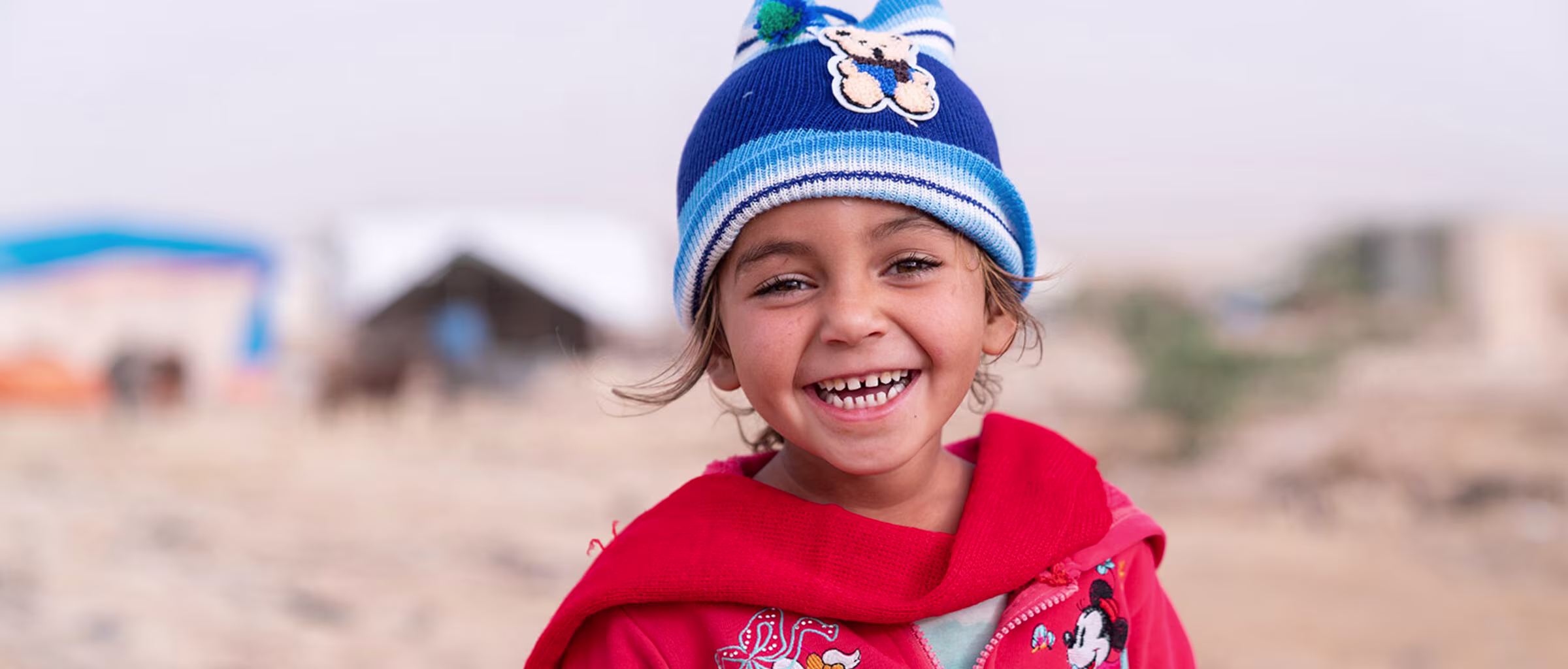 A smiling child wearing a blue hat and a red hoodie in a desert-like setting with buildings in the background.