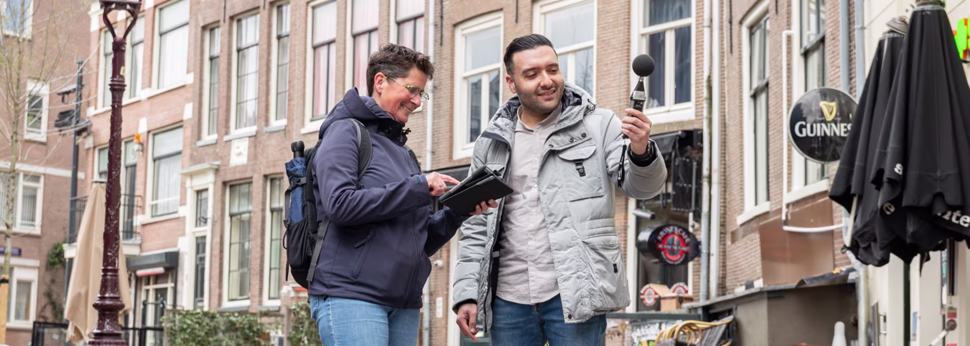 Twee mannen voeren een vriendelijk gesprek op straat, één met klembord en één met microfoon, met historische gebouwen op achtergrond.