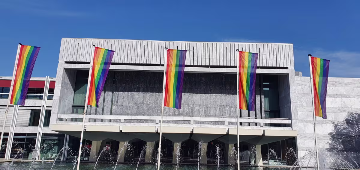 Modern gemeentehuis van Arnhem met regenboogvlaggen en fontein, onder blauwe hemel.