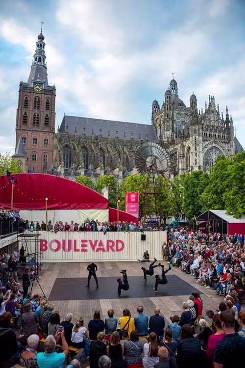 Outdoor theatervoorstelling op een plein met publiek rond het podium, op de achtergrond een grote gotische kerk.