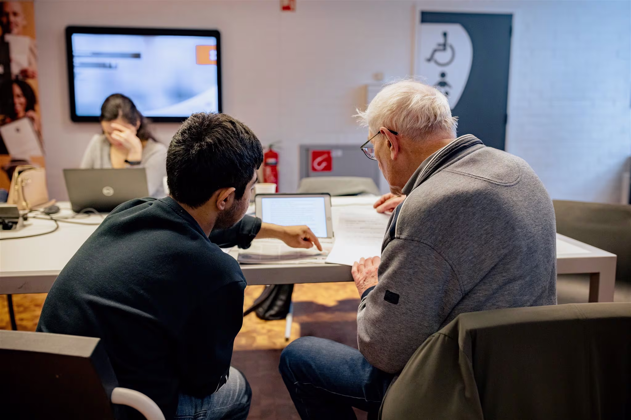 Een medewerker helpt een oudere man bij een laptop in een moderne bibliotheek met witte tafels en stoelen.