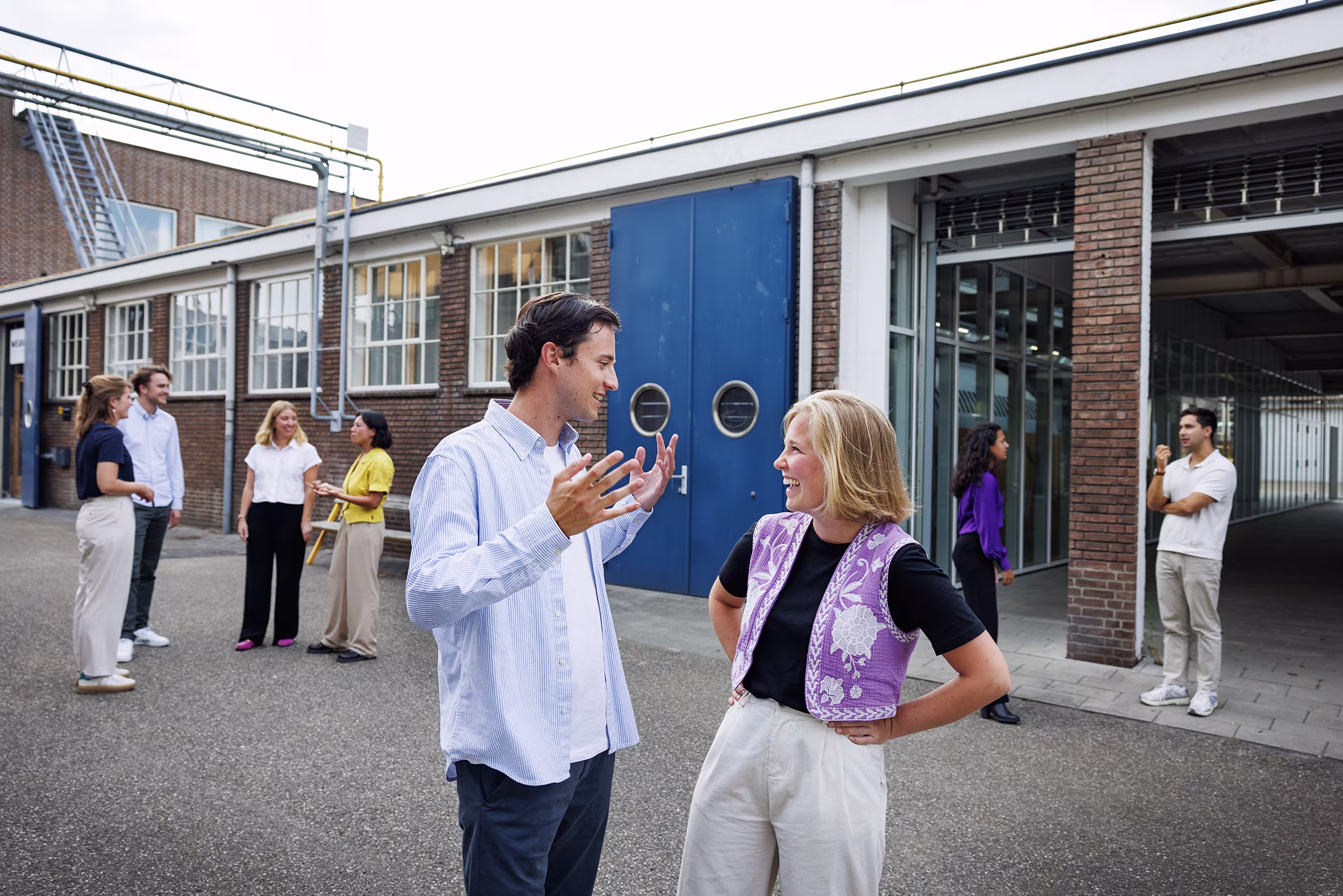 A man in a blue shirt is talking enthusiastically with a blonde woman in a purple cardigan; in the background are several other colleagues.