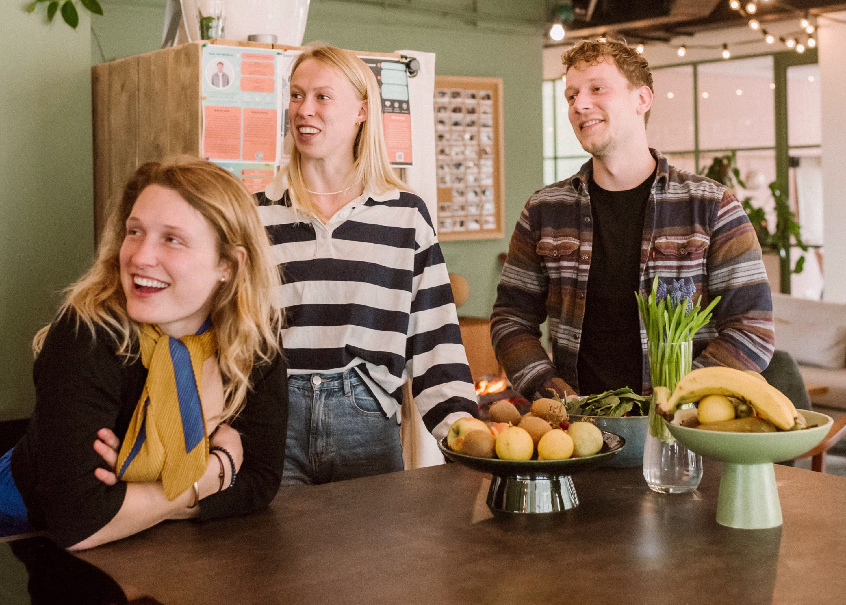 Three smiling coworkers are standing around a table with fruit in a modern office setting with green walls and string lights.