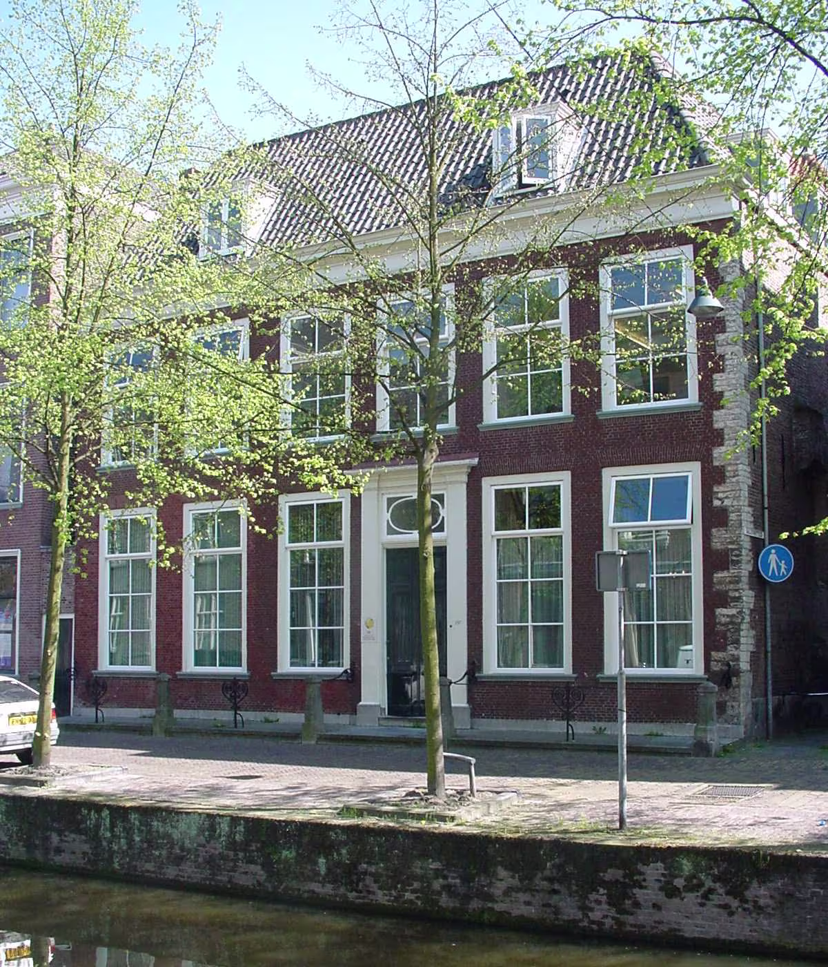 Historisch bakstenen kantoorpand aan de gracht met grote witte ramen en pannendak, omgeven door groene bomen.