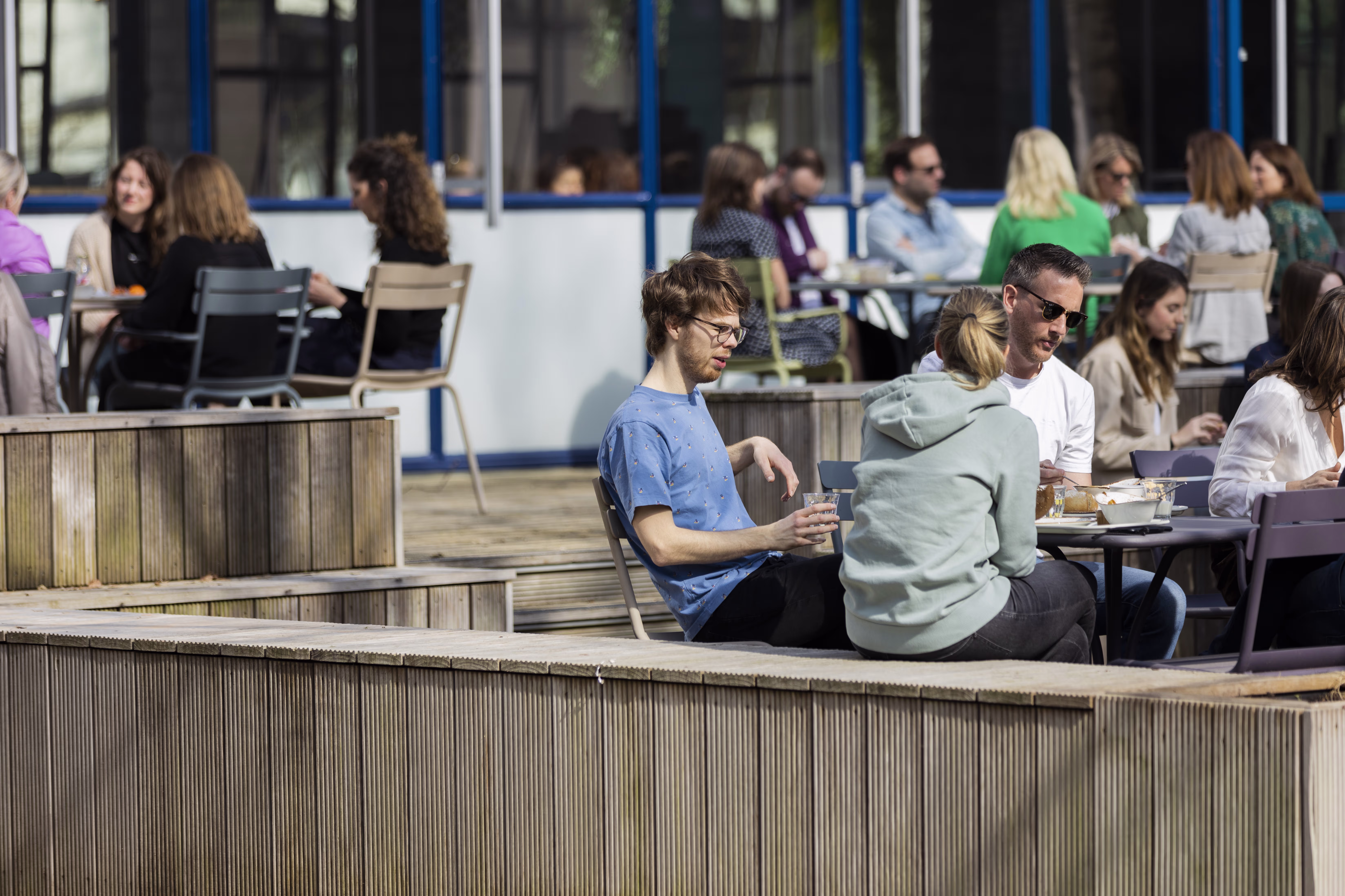 Mensen zitten ontspannen op houten terrassen en tafels in de zon, pratend en etend in een moderne buitenruimte.