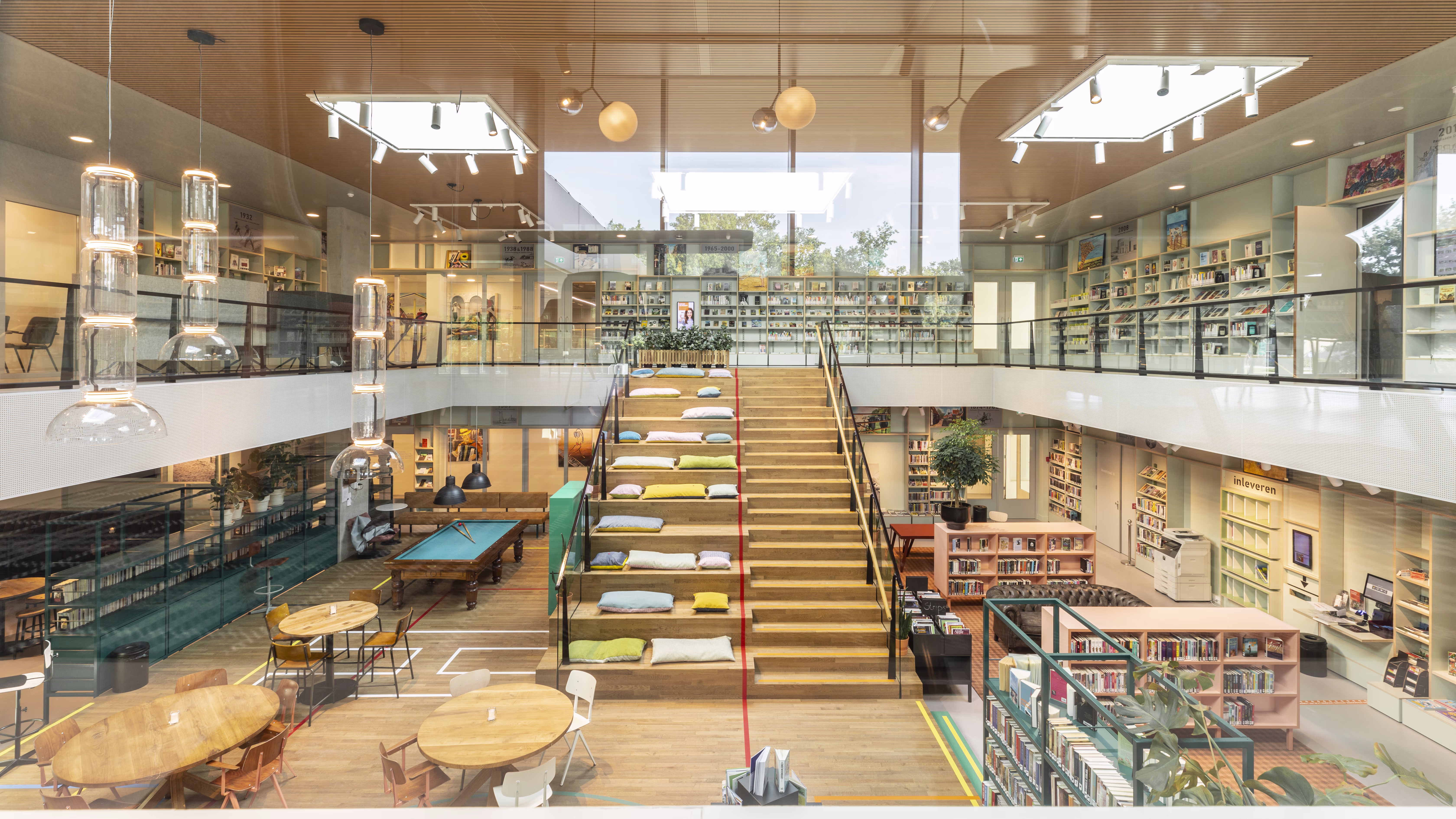 Moderne bibliotheek met glazen trap, boekenkasten op twee verdiepingen, zitplekken en natuurlijk licht door grote ramen.
