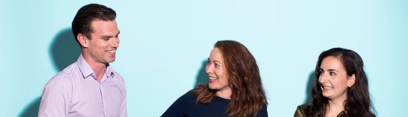 Drie lachende collega's in gesprek tegen een lichtblauwe achtergrond, een man in lila overhemd en twee vrouwen.
