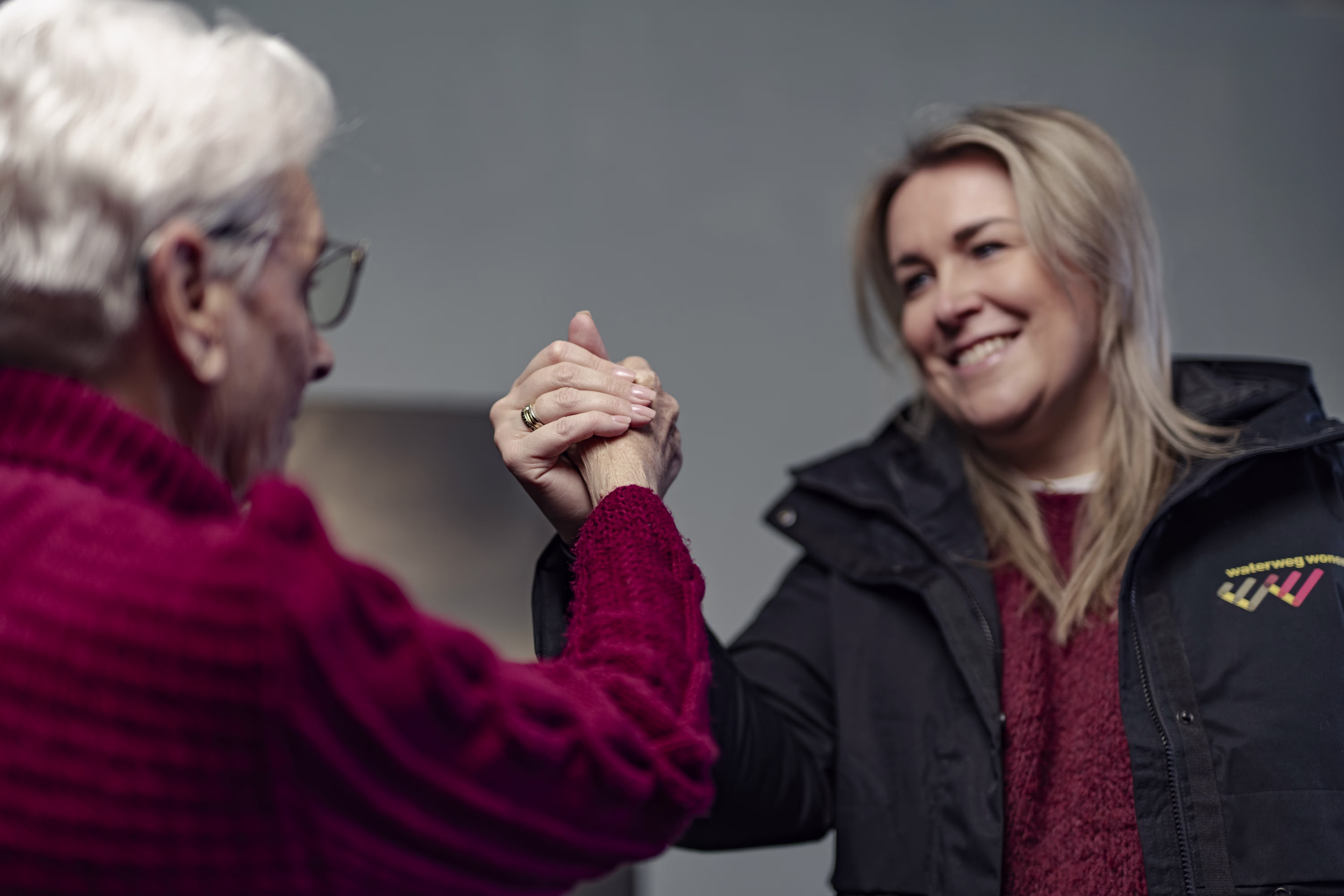 Een oudere vrouw en jongere blonde vrouw schudden elkaar lachend de hand, beide dragen rode truien.