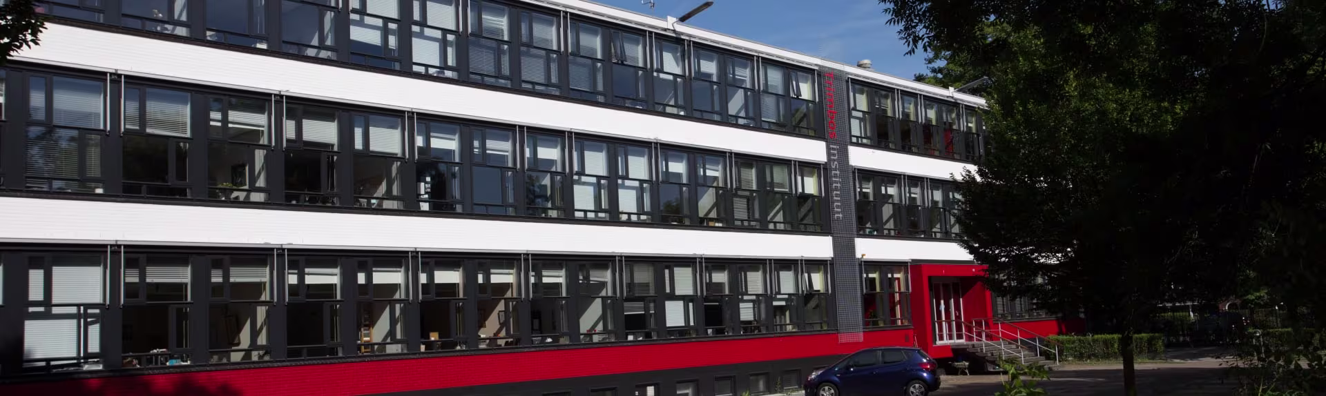 Buitenkant van een modern kantoorgebouw met rode accenten en grote ramen, omgeven door bomen.