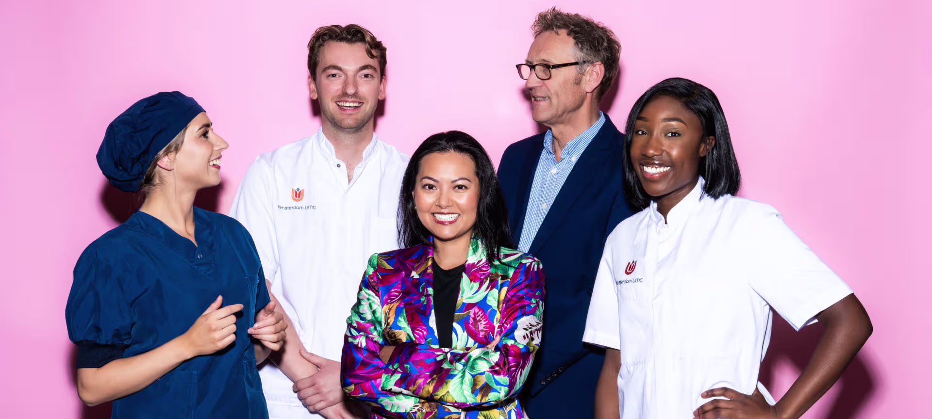 Vijf lachende medewerkers van Amsterdam UMC poseren samen voor een roze achtergrond, in medische en zakelijke kleding.