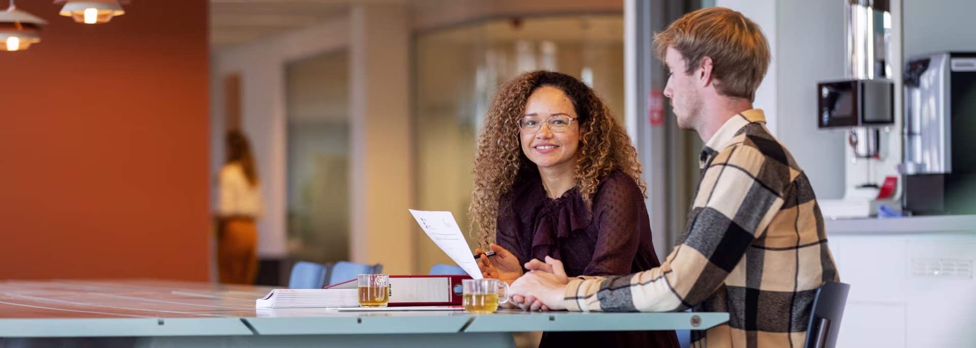 Twee collega's bespreken documenten aan een tafel in een modern kantoor, met glazen thee voor hen.