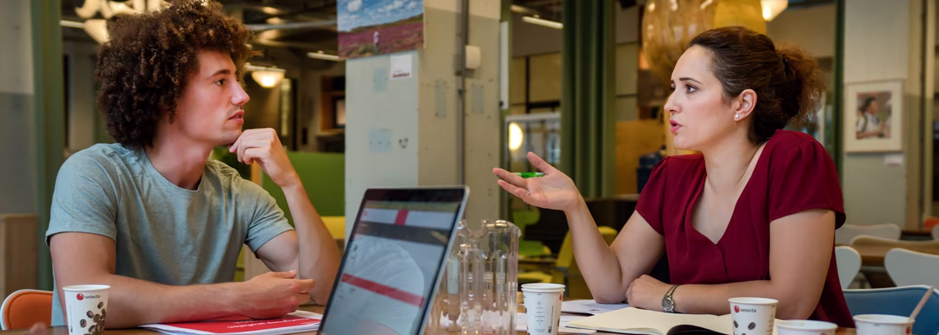 Twee collega's in gesprek aan een werktafel met laptop en koffiebekers in een moderne kantooromgeving.