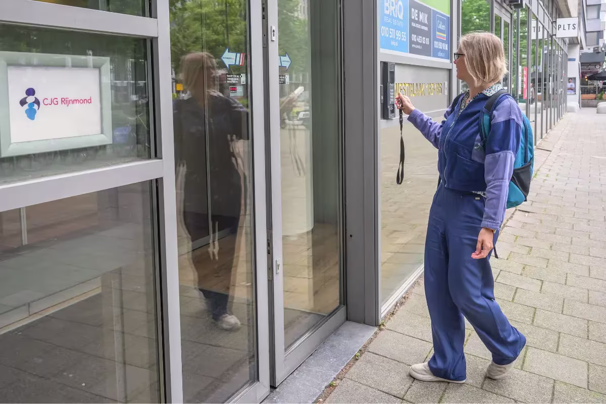 Een vrouw in blauwe jumpsuit belt aan bij een glazen gebouw met het CJG Rijnmond-logo.