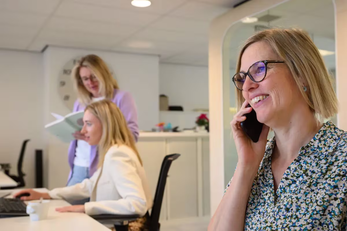 Drie vrouwen werken samen op kantoor; één belt, één werkt aan een computer, één bekijkt een document.