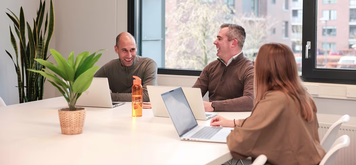 Drie collega's overleggen samen aan een vergadertafel met laptops in een lichte, moderne kantoorruimte.