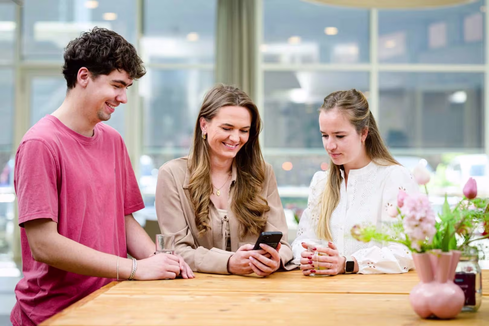 Three colleagues are looking at a smartphone together at a wooden table in a bright, modern office space.
