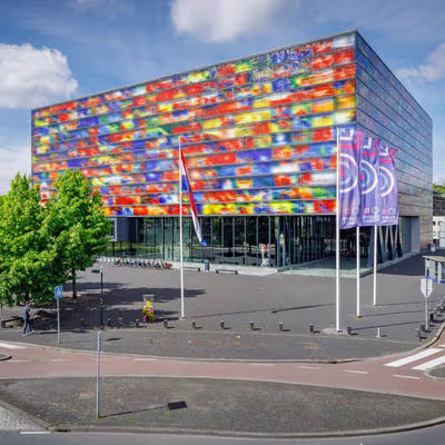 Kleurrijk glazen gebouw van Beeld & Geluid in Hilversum, met vlaggen en bomen op een zonnige dag.