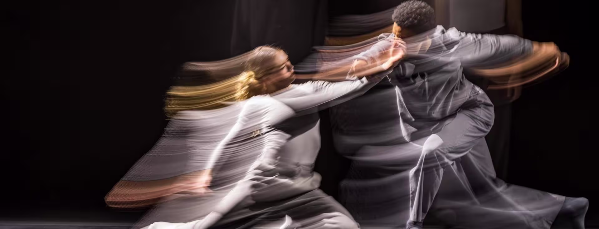 Twee dansers in grijze kleding bewegen dynamisch samen, vastgelegd met motion blur op een donkere achtergrond.