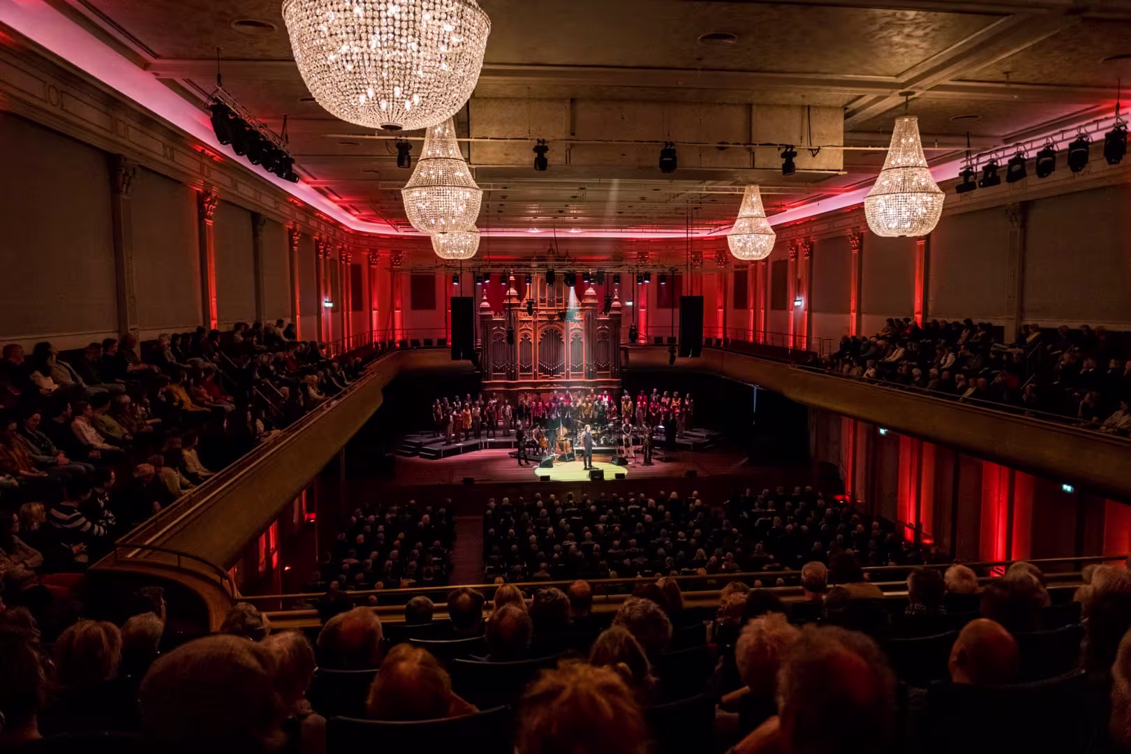 Volle concertzaal met rood licht, kroonluchters en een koor op het podium voor een groot orgel.
