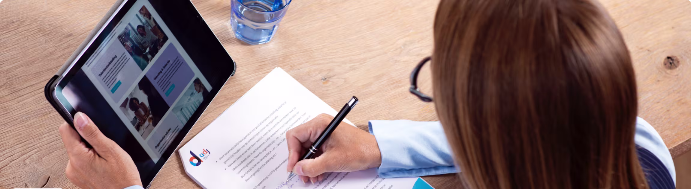 Vrouw schrijft op papier met logo terwijl ze een tablet vasthoudt met daarop een website.