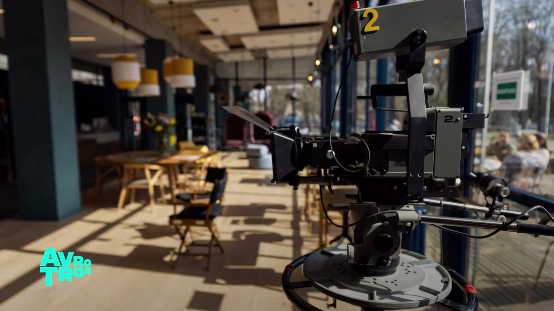 Een professionele tv-camera met nummer 2 op een filmset in een lichte studio met tafels en stoelen.