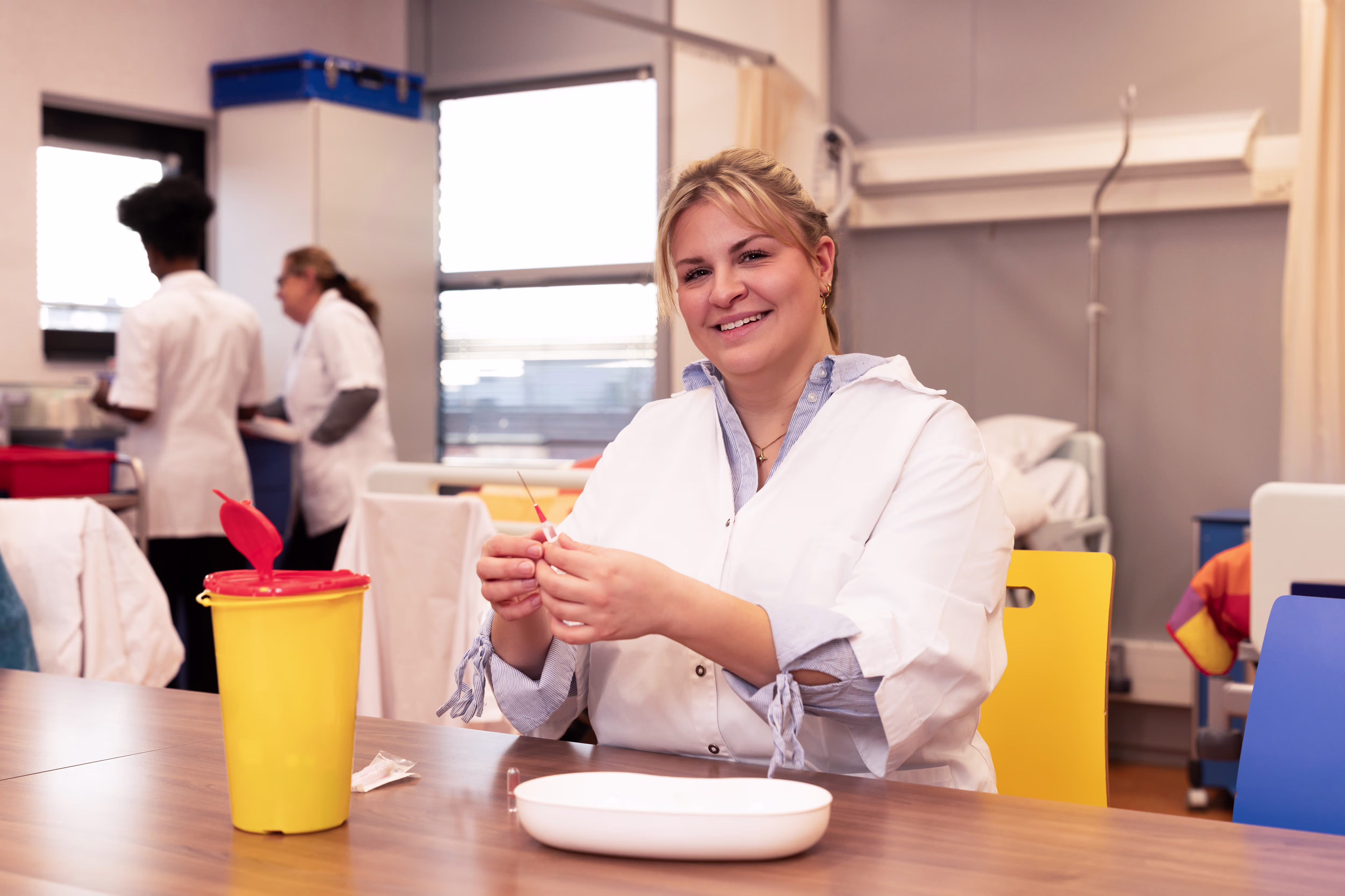Verpleegkundestudent in witte jas oefent met naald bij een sharps container in een vaardigheidslokaal.