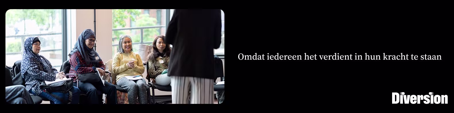 Vier vrouwen zitten in een kring en luisteren aandachtig naar een staande spreker tijdens een bijeenkomst.