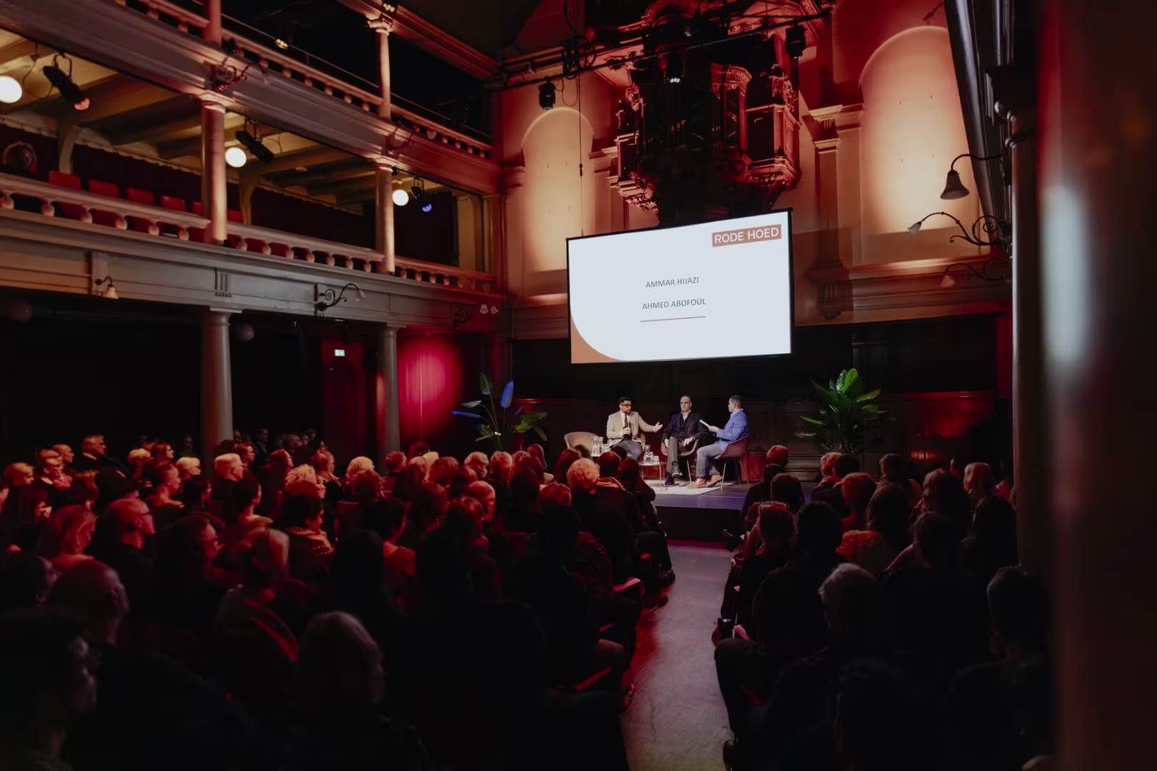 Volle zaal bij een panelgesprek op het podium van de Rode Hoed in Amsterdam, met publiek en sprekers op het verlichte podium.