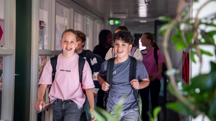Twee lachende leerlingen met rugzakken lopen door een schoolgang, gevolgd door klasgenoten.