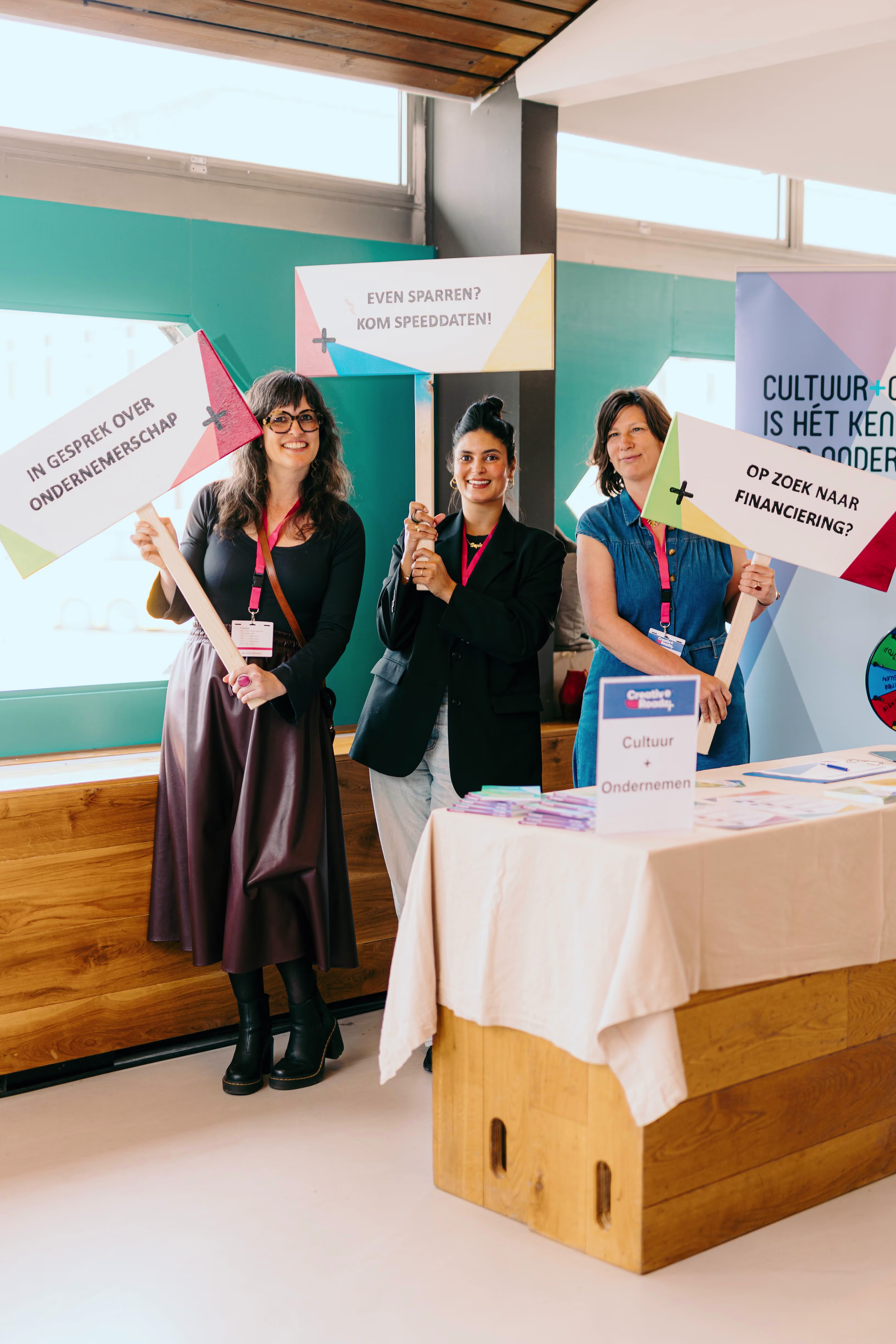 Three smiling women holding signs about culture and entrepreneurship at an information table at the Stedelijk.