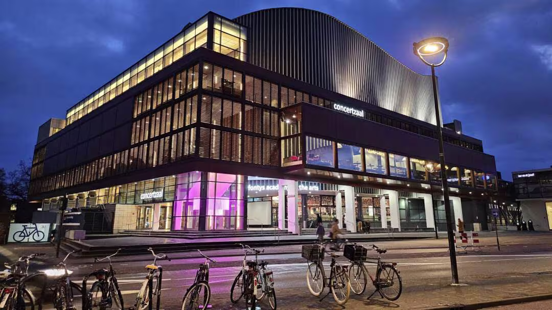 Concertzaal en Fontys Academie for the Arts, verlicht in de avond, met fietsen op de voorgrond.