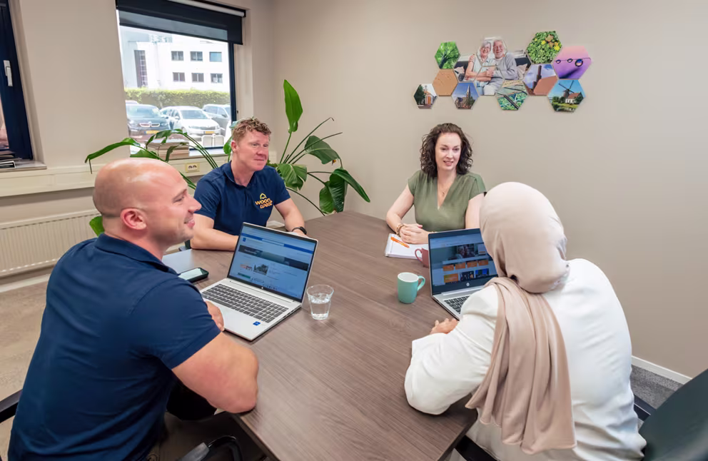 Vier collega's van WoonWest overleggen aan een vergadertafel met laptops in een lichte vergaderruimte.