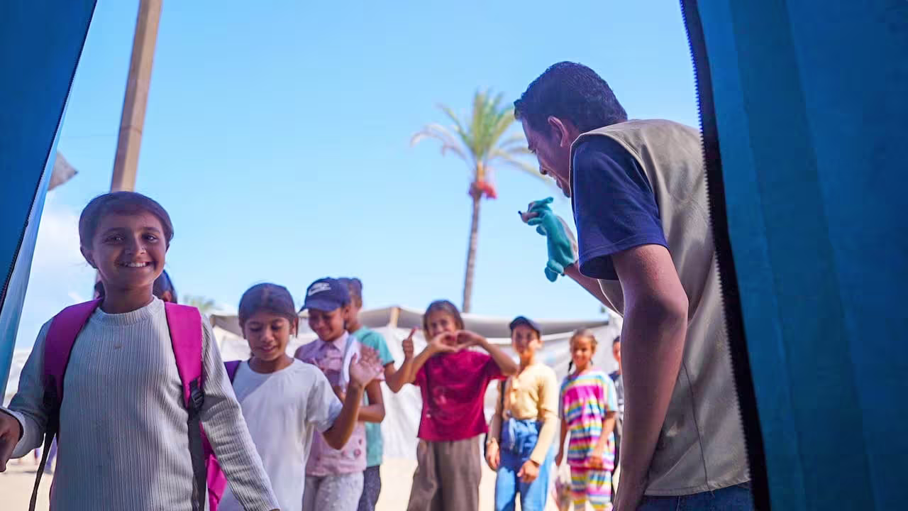 A staff member holding a hand puppet welcomes smiling children at a blue tent, outside under a blue sky.