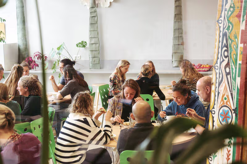 Medewerkers lunchen samen aan lange tafels in een kleurrijke, gezellige kantine met groene stoelen en planten.