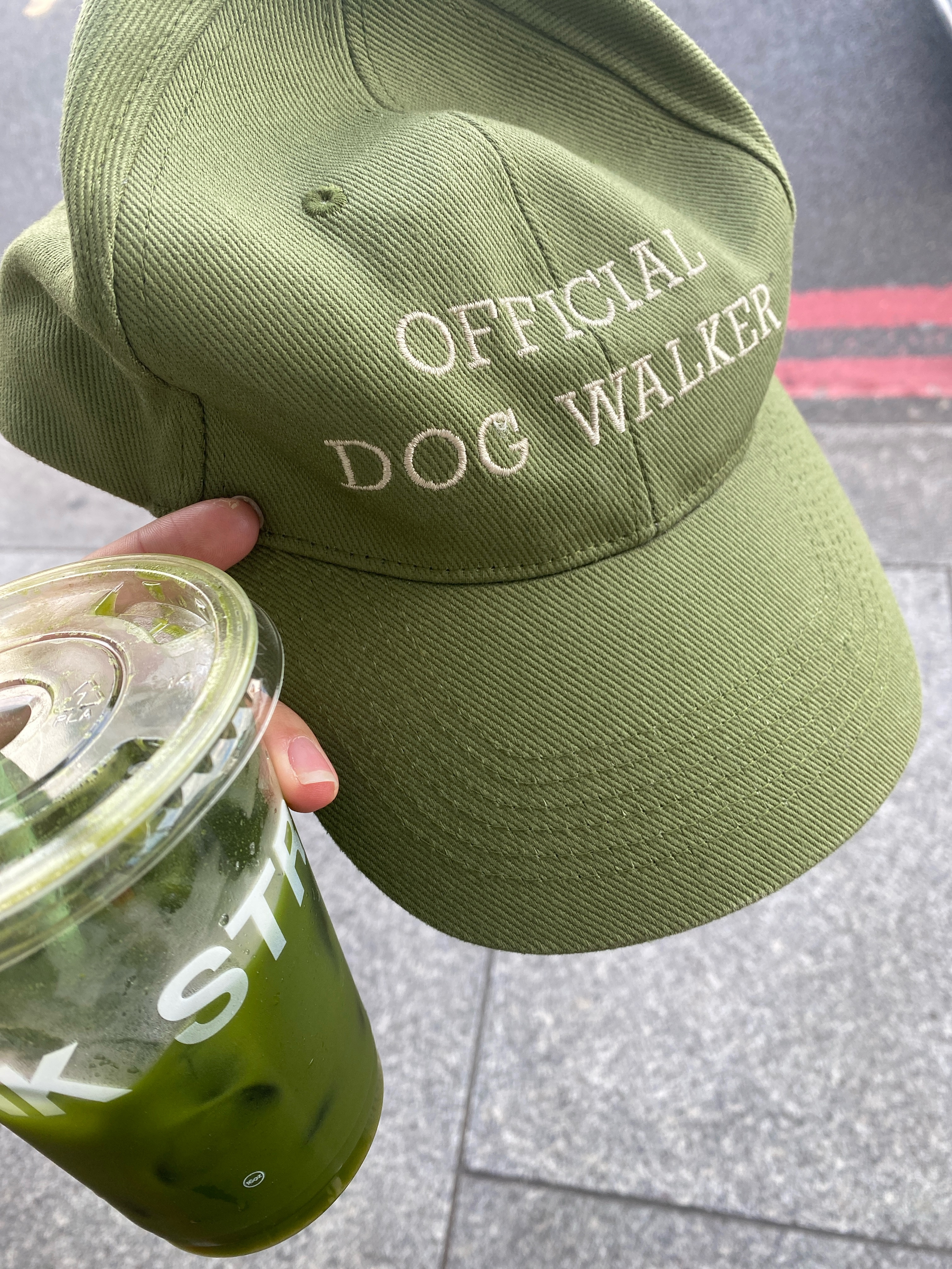 Close-up shot of someone holding a drink and a green cap that says &ldquo;OFFICIAL DOG WALKER&rdquo; in all caps