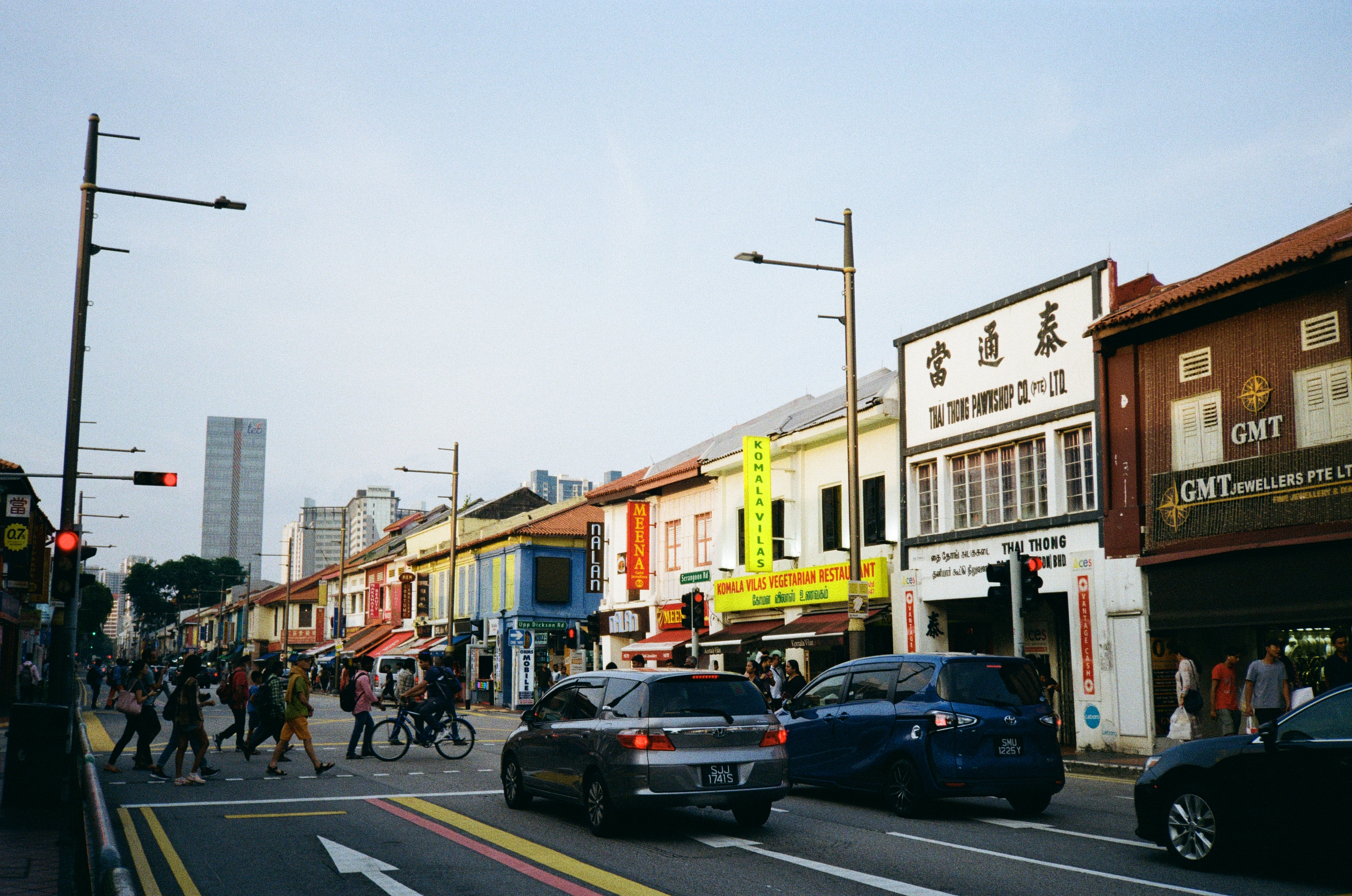 Stretch of shops along Serangoon Rd, around the late afternoon. There are pedestrians crossing the road. Both sides of the road are flanked by shophouses.