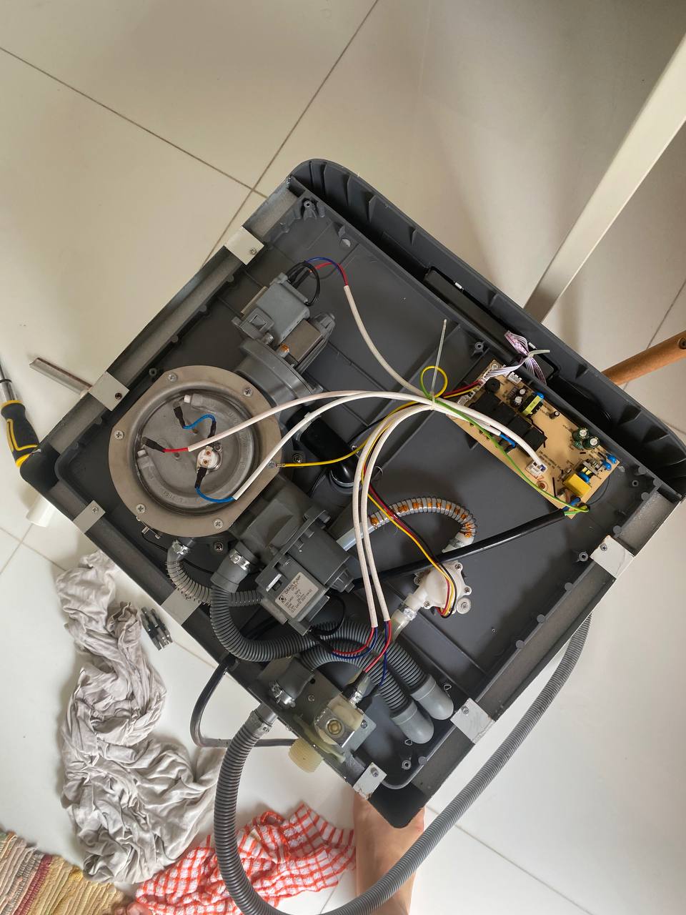 Underside of a dishwasher with internals exposed. There is a heating element, an inlet and draining hose, and a small circuit.