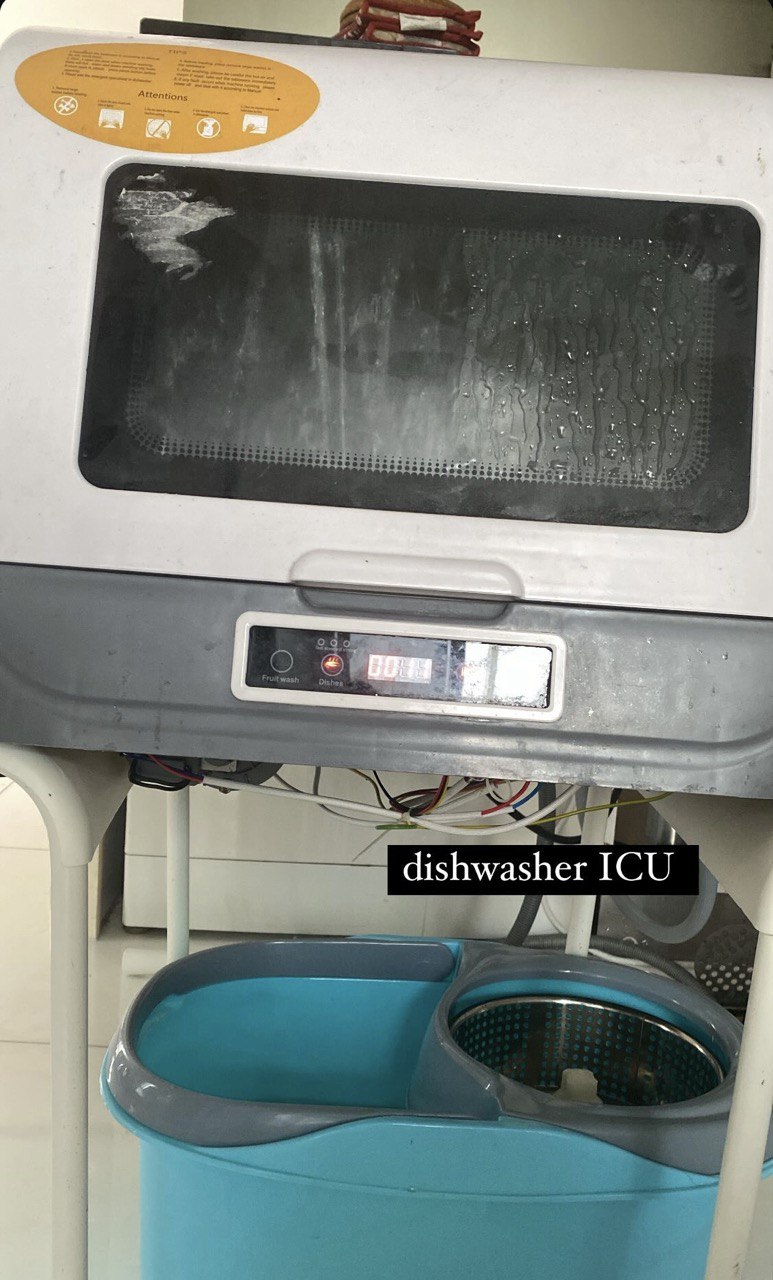Dishwasher propped up with 2 chairs on either side, with a blue mop bucket directly underneath it