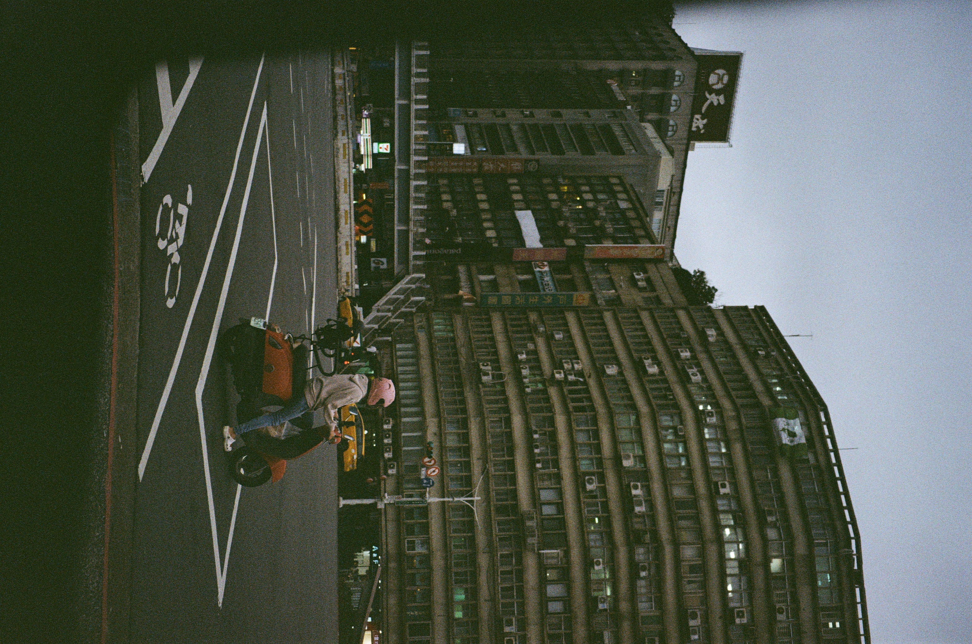 The sky is overcast. A person riding a red scooter is in the foreground at a traffic junction while the background comprises of a large building and an overhead pedestrian bridge.
