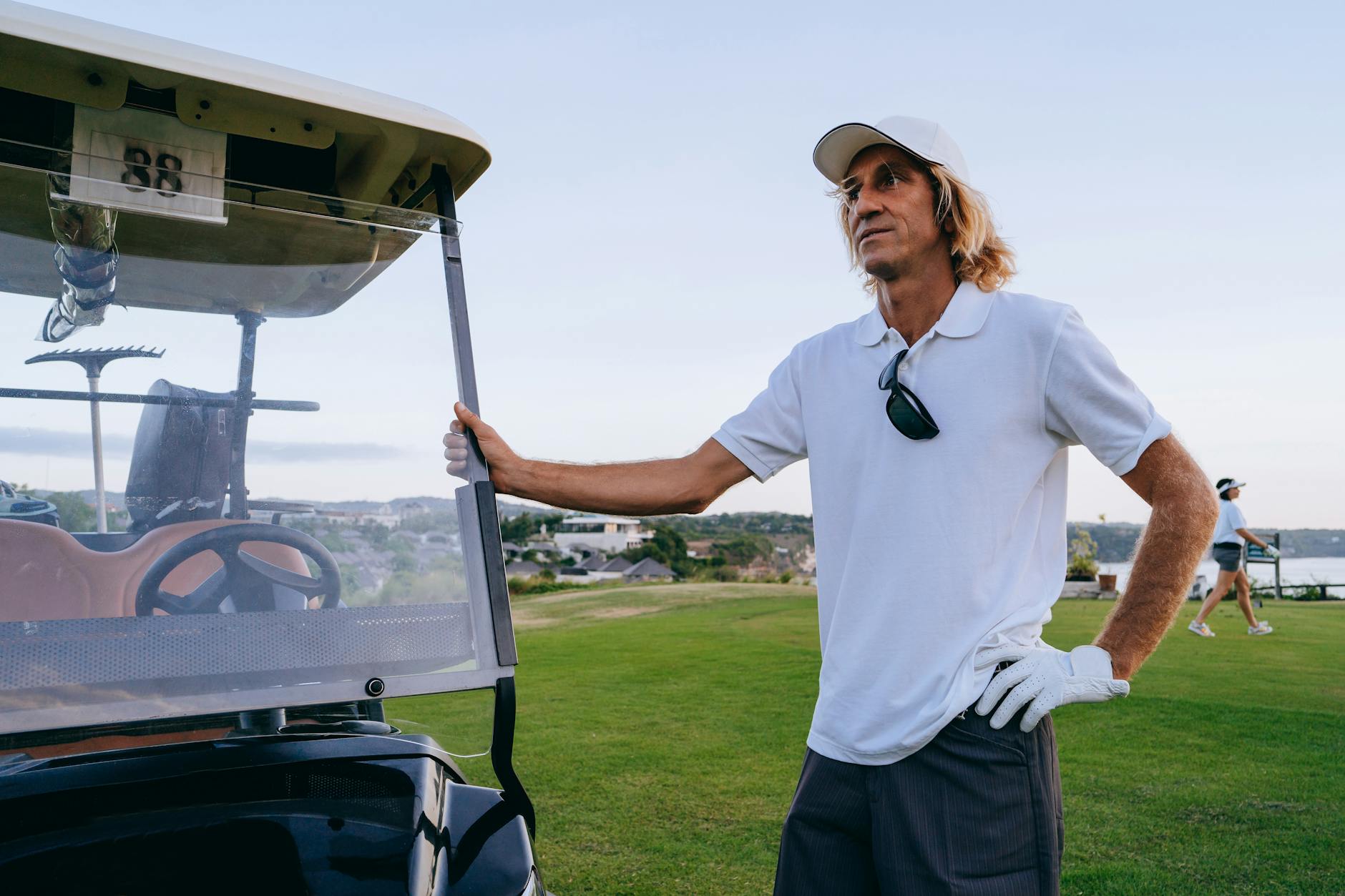 A man in a white shirt stands by a golf cart on a lush golf course during the day.
