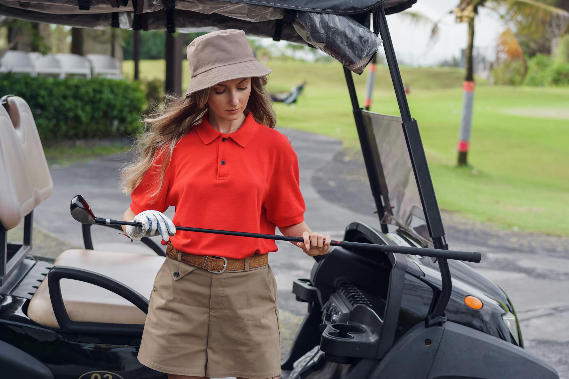Woman in red and khaki golfing outfit standing by a cart, holding a golf club on a sunny day.