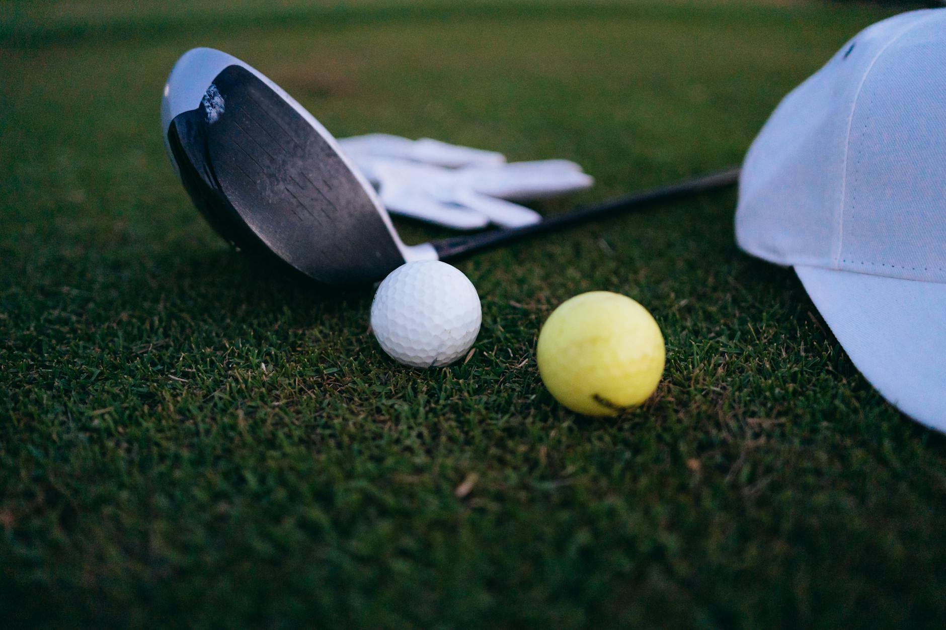 Close-up shot of a golf club, golf balls, and a cap on grass.
