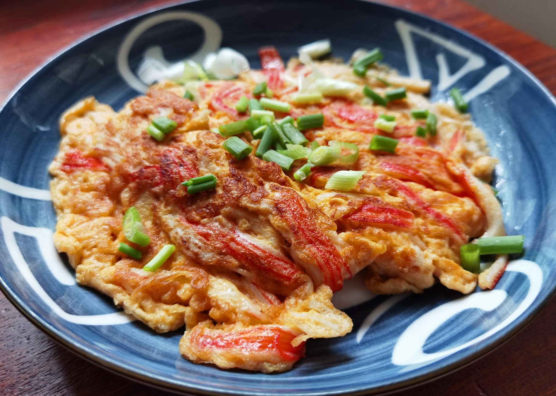 Close-up of a savory crab omelet garnished with fresh spring onions, served on a decorative ceramic plate.