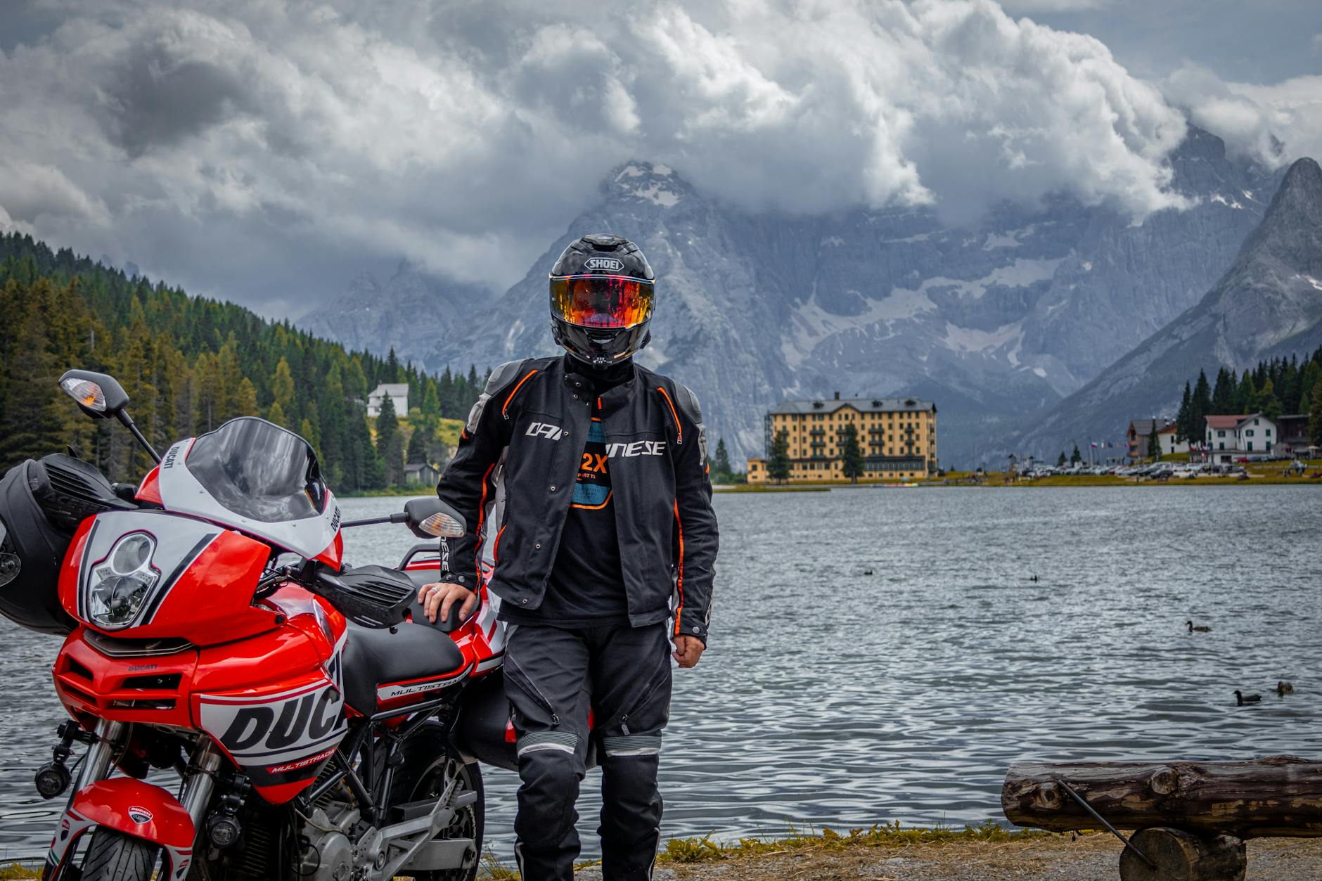A motorcyclist poses by a scenic alpine lake surrounded by dramatic mountains and a historic building.
