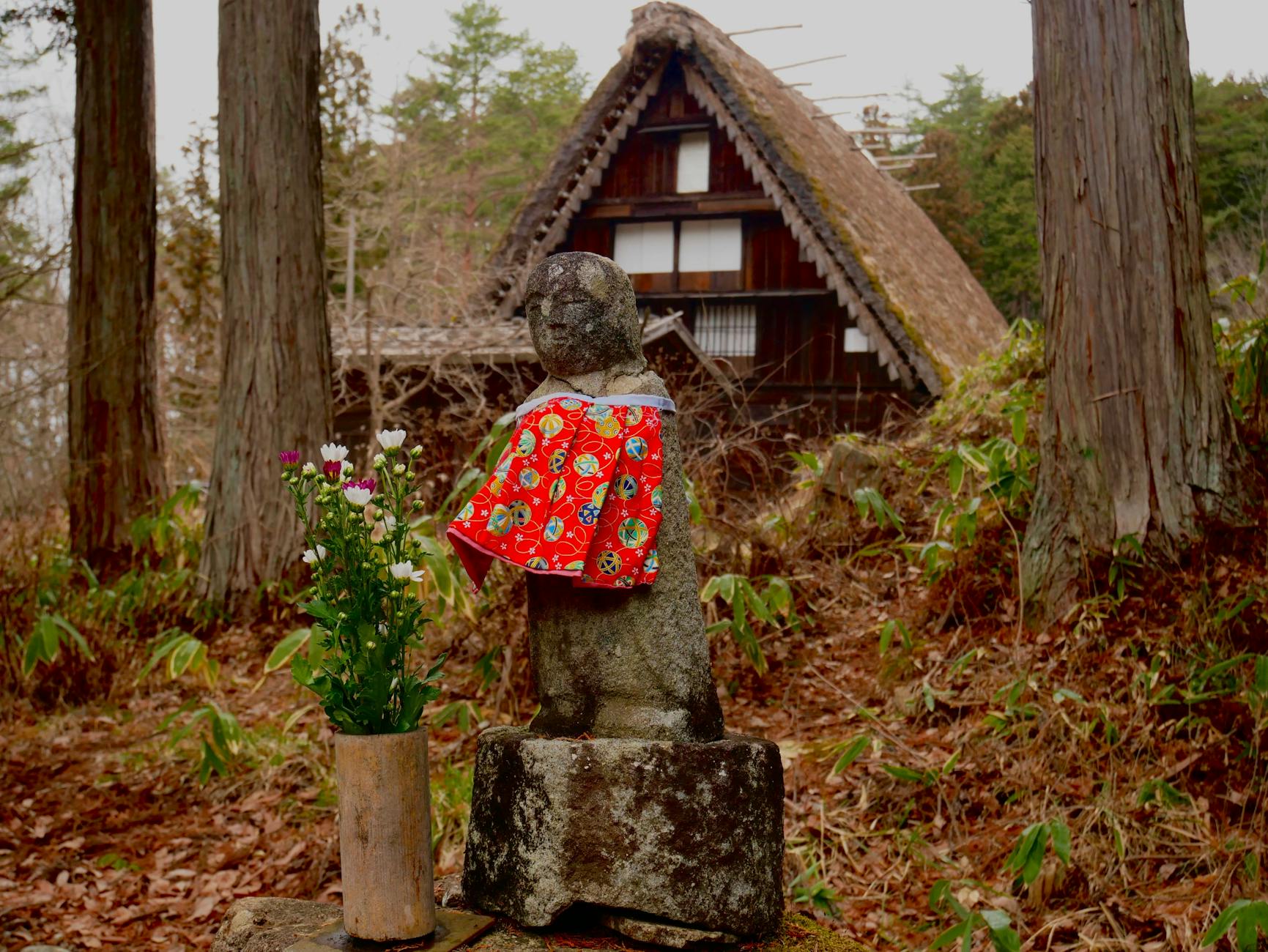 Stone statue wearing traditional clothing in Takayama garden with flowers and thatched roof house.