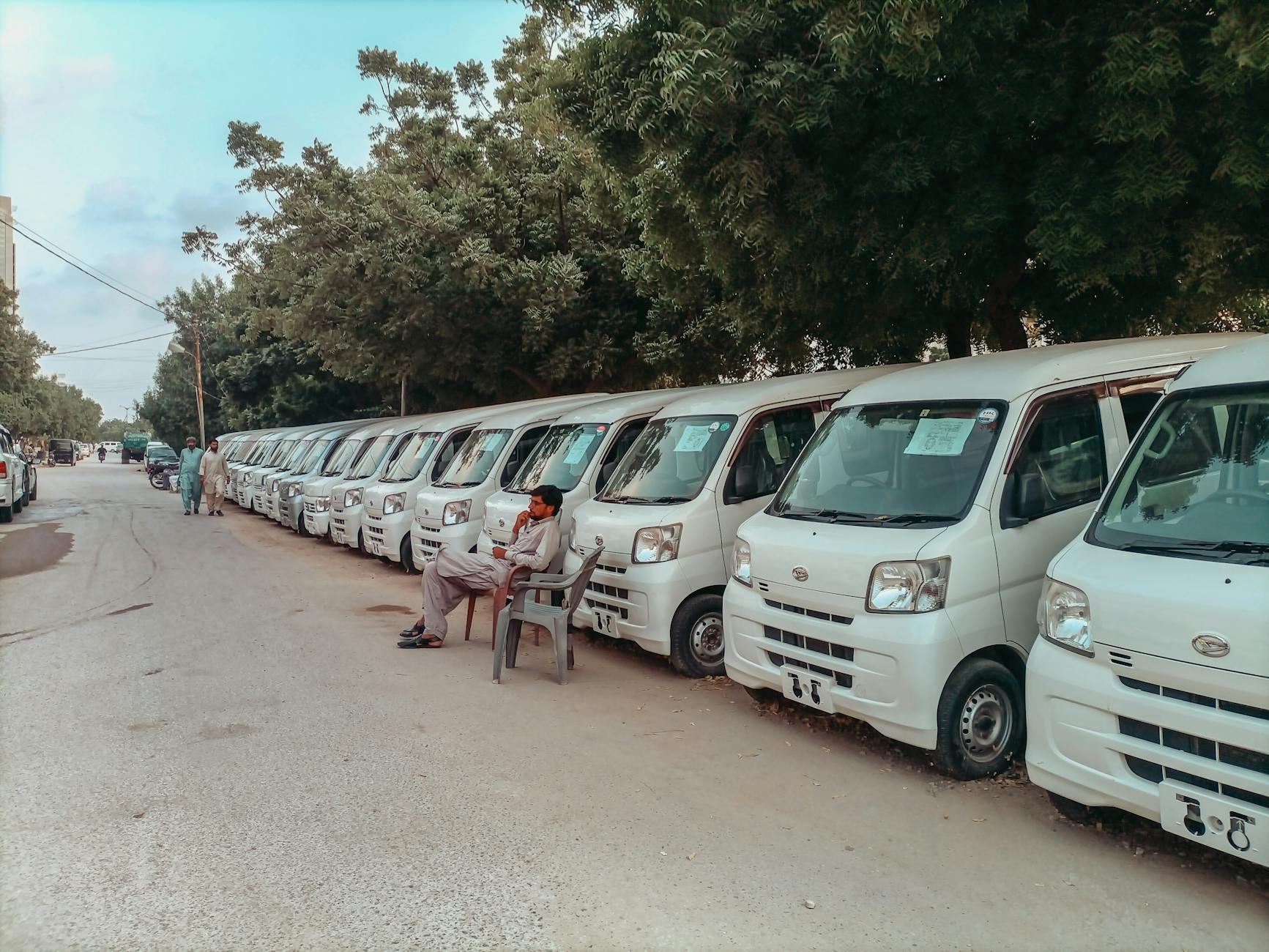 Row of white vans lined up on a street with people, trees, and relaxed atmosphere.