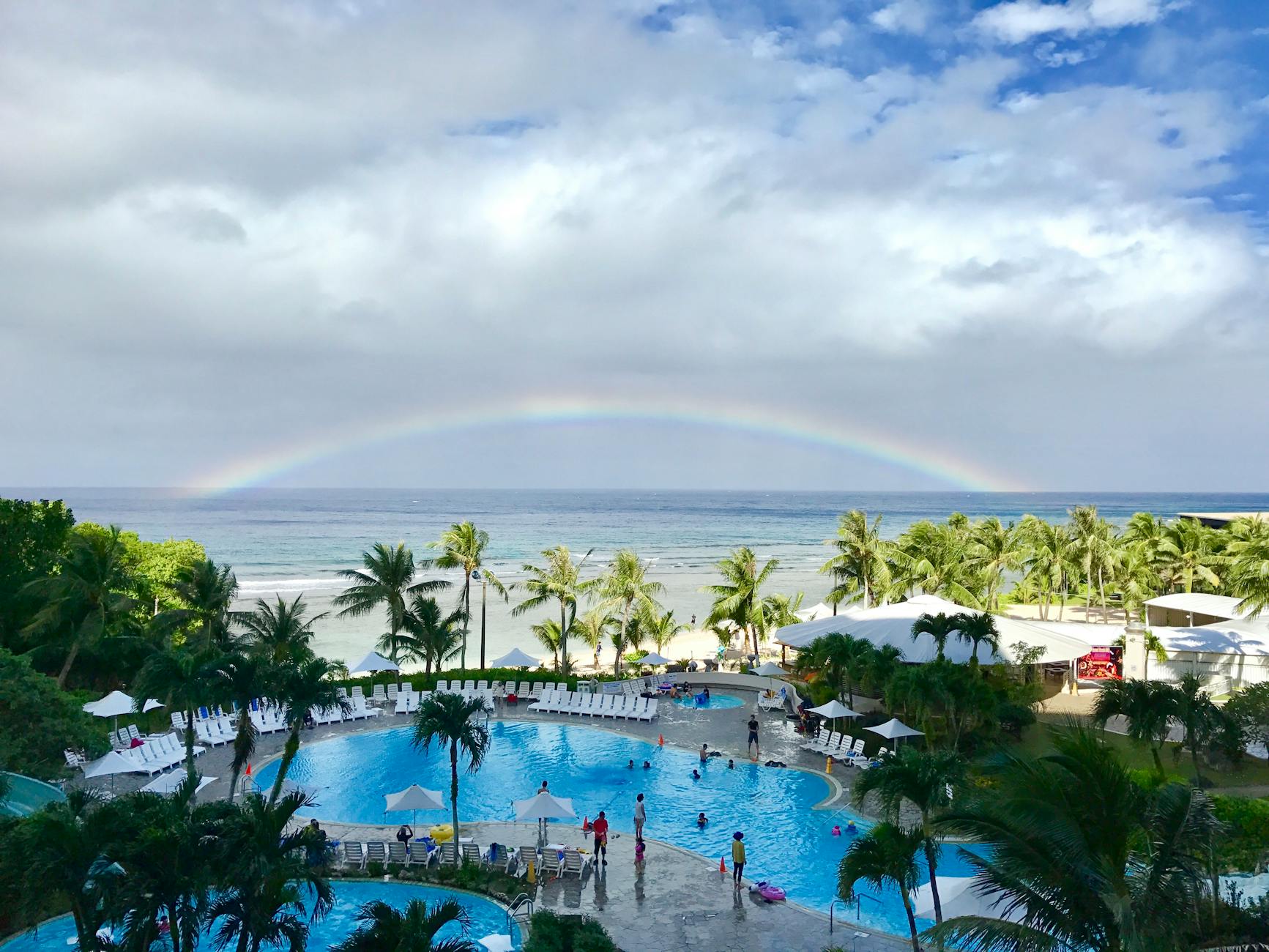 Aerial view of a tropical resort pool in Tumon, Guam with a vibrant rainbow above the ocean, showcasing palm trees and a summer ambiance.