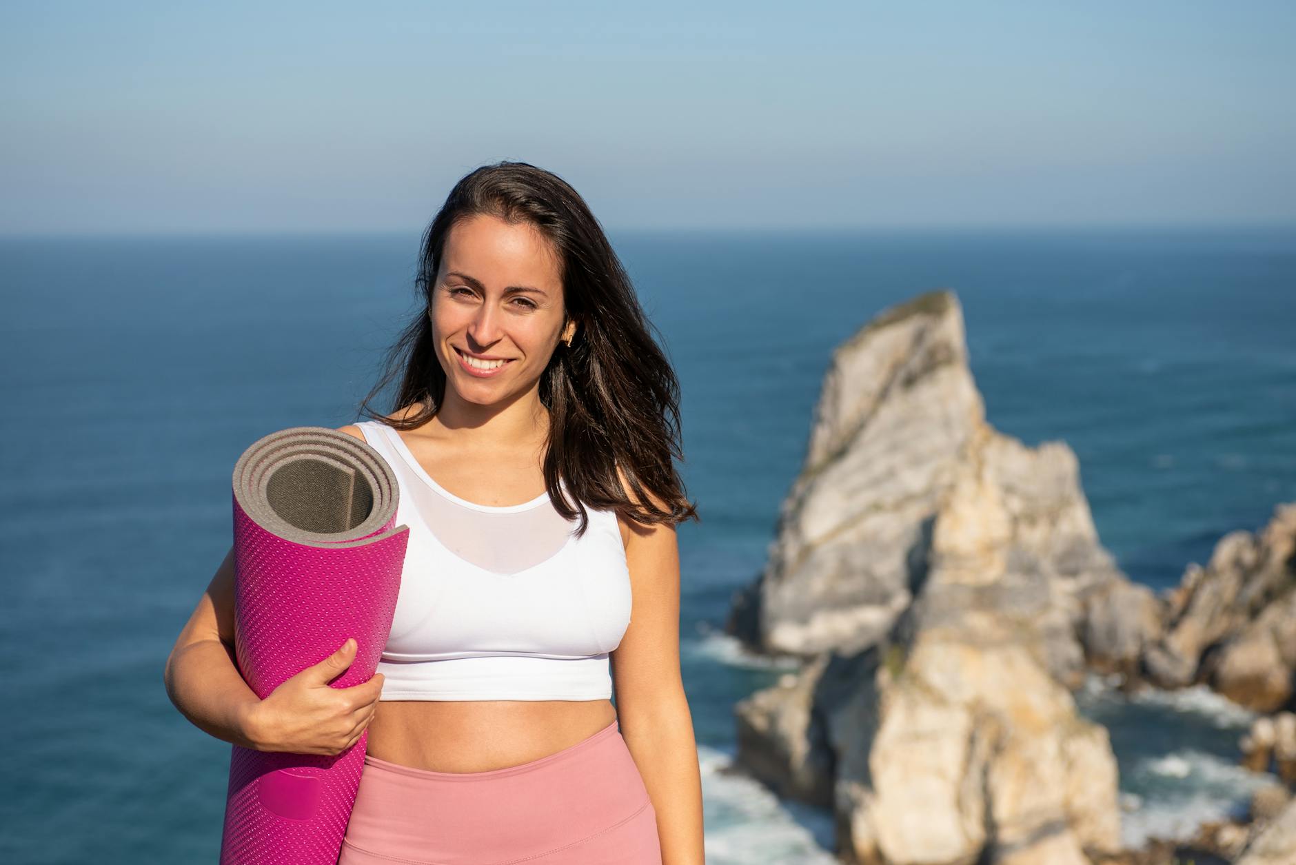Smiling woman with yoga mat in sportswear by oceanic cliffs in Portugal.