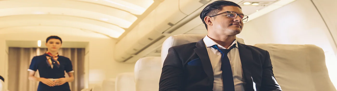 A man in a formal suit sitting comfortably on an airplane seat, while a flight attendant stands in the aisle, symbolizing Turkish Airlines’ in-flight comfort and professional service.